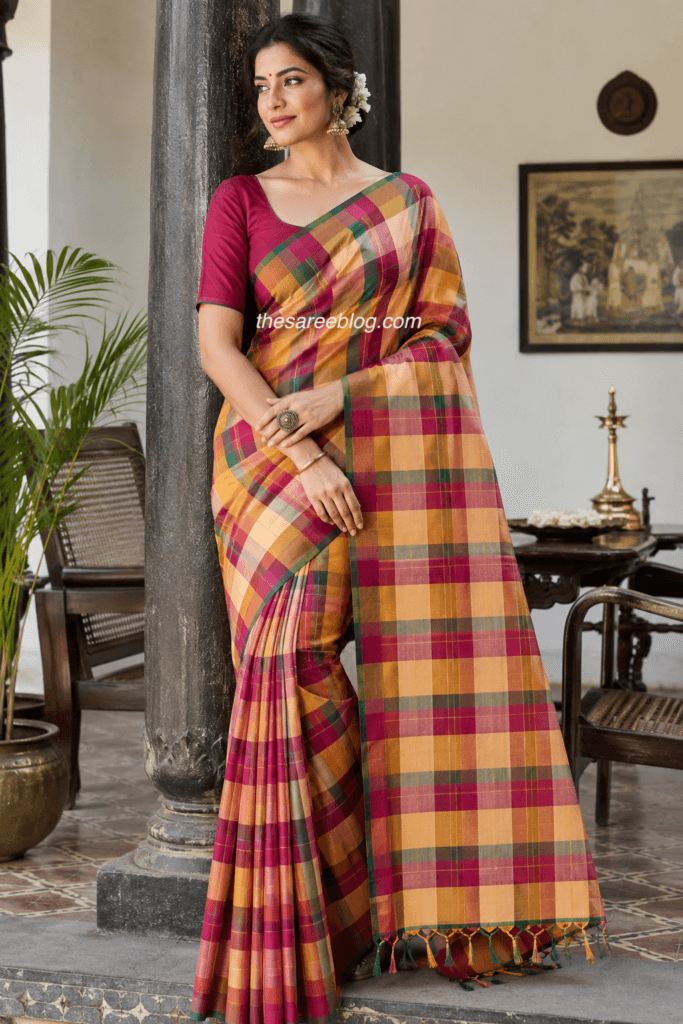 Woman wearing a traditional Madras Check cotton saree in vibrant mustard, magenta, and green plaid, paired with a magenta blouse, gold jhumkas, and jasmine flowers, standing in a heritage South Indian courtyard.