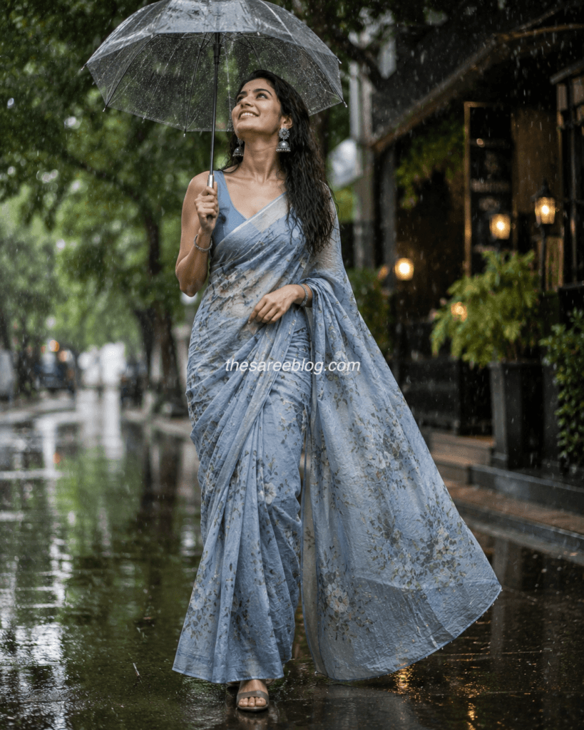 Woman wearing a pastel blue floral saree with a matching blouse, walking through a rain-soaked street under a transparent umbrella, showcasing elegant monsoon saree styling.